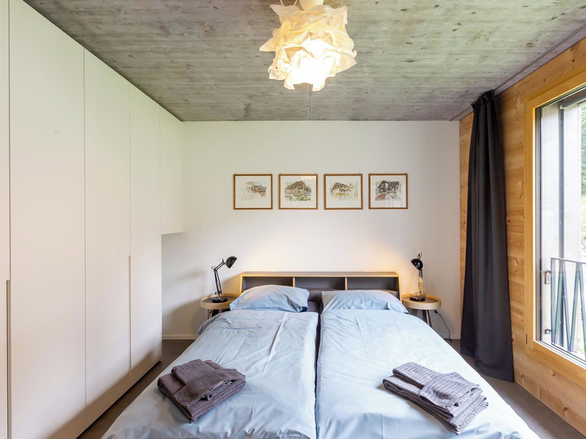 Un dormitorio con una cama doble, dos mesitas de noche con lámparas, un candelabro en el techo y cuatro cuadros enmarcados en la pared.