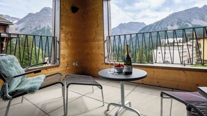 Een balkon met uitzicht op bergen, twee stoelen, een tafel met een fles en glazen, en een houten wand.