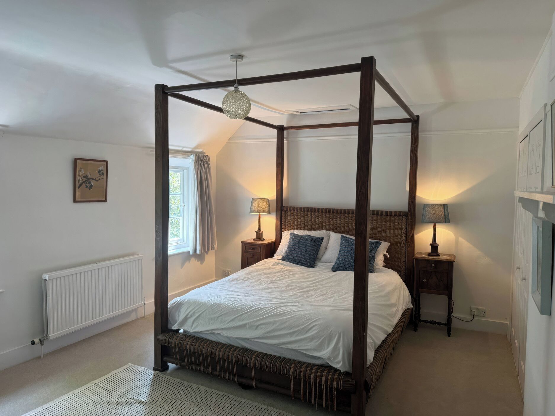 Un lit à baldaquin, deux tables de chevet avec des lampes, un radiateur, une fenêtre avec des rideaux et un tableau encadré au mur.