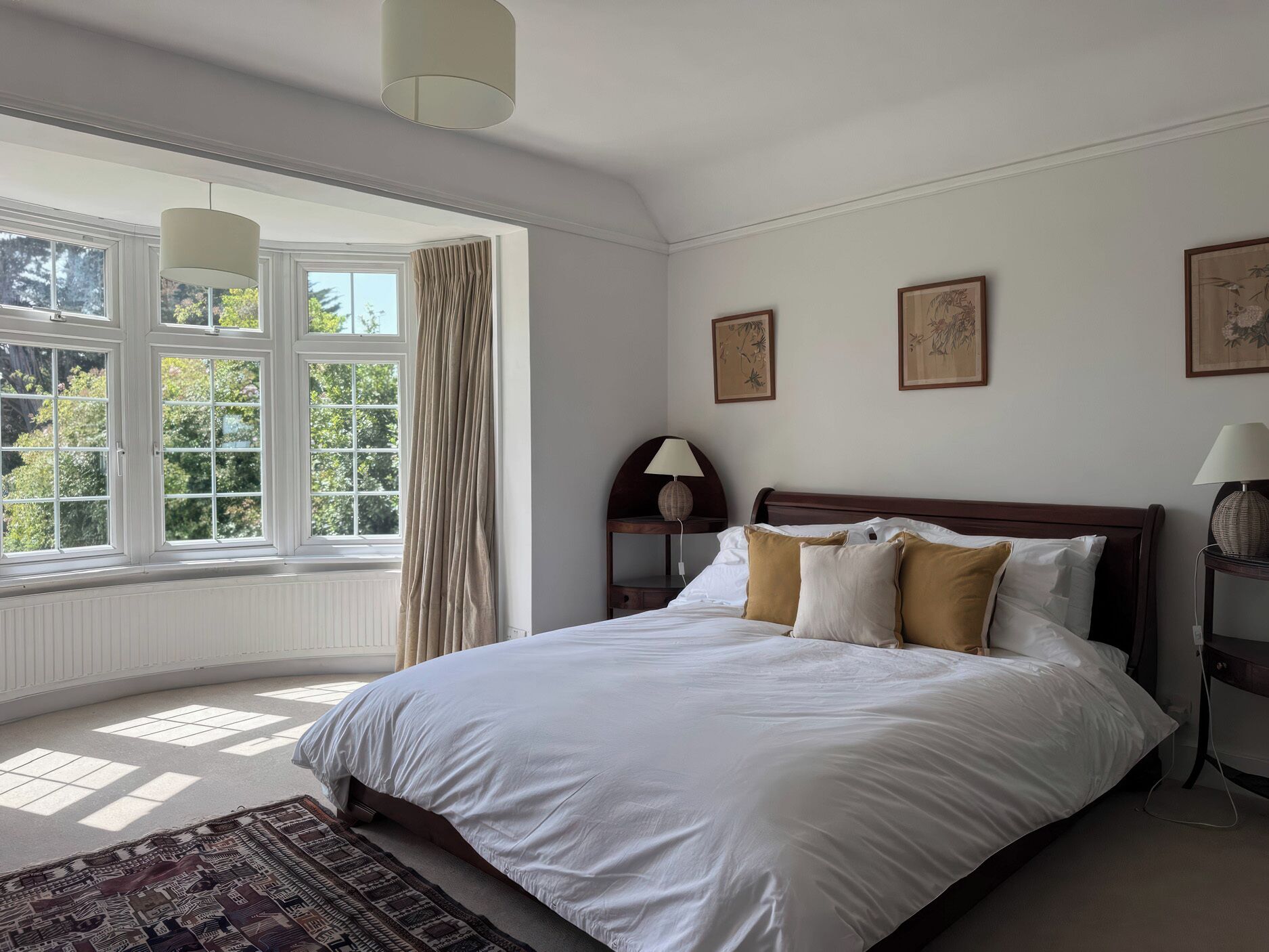 Une chambre avec une grande fenêtre, un lit avec une literie blanche et marron, une table de chevet avec une lampe, et un tapis sur le sol.