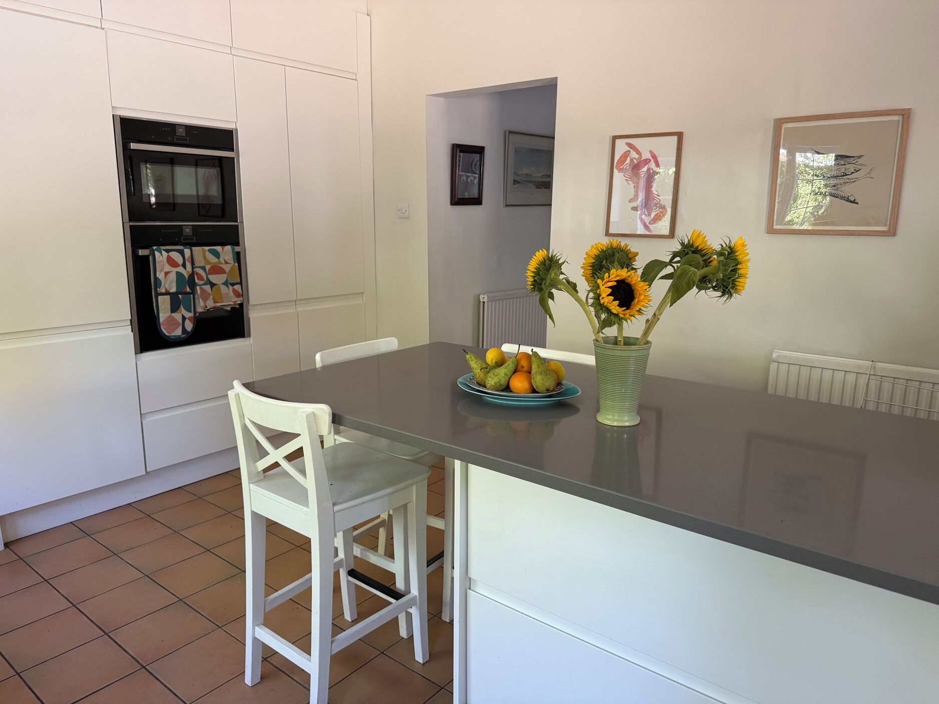 Una cocina con armarios blancos, horno empotrado, una mesa de comedor con un jarrón de girasoles y un plato de fruta.