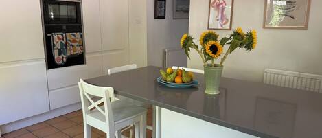 Una cocina con armarios blancos, horno empotrado, una mesa de comedor con un jarrón de girasoles y un plato de fruta.