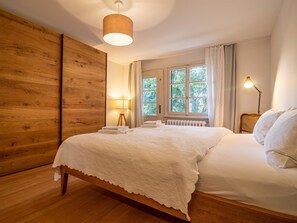 Une chambre avec une armoire en bois, un lit avec du linge de lit blanc, une fenêtre avec des rideaux et une lampe de chevet.