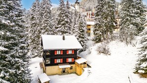 Ein mit Schnee bedecktes Gebäude mit einem steilen Dach, umgeben von schneegefüllten Bäumen und einem in der Ferne sichtbaren Kirchturm.