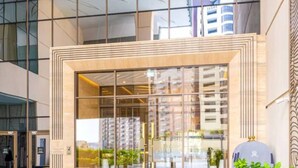 Entrada de um edifício moderno com portas de vidro, estrutura metálica e um pequeno jardim com plantas.