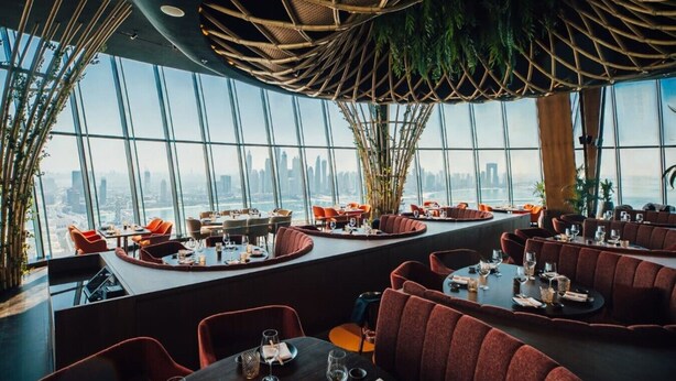 Um restaurante com mesas redondas e bancos estofados vermelhos, amplas janelas com vista para a cidade e um design de teto em tecido.
