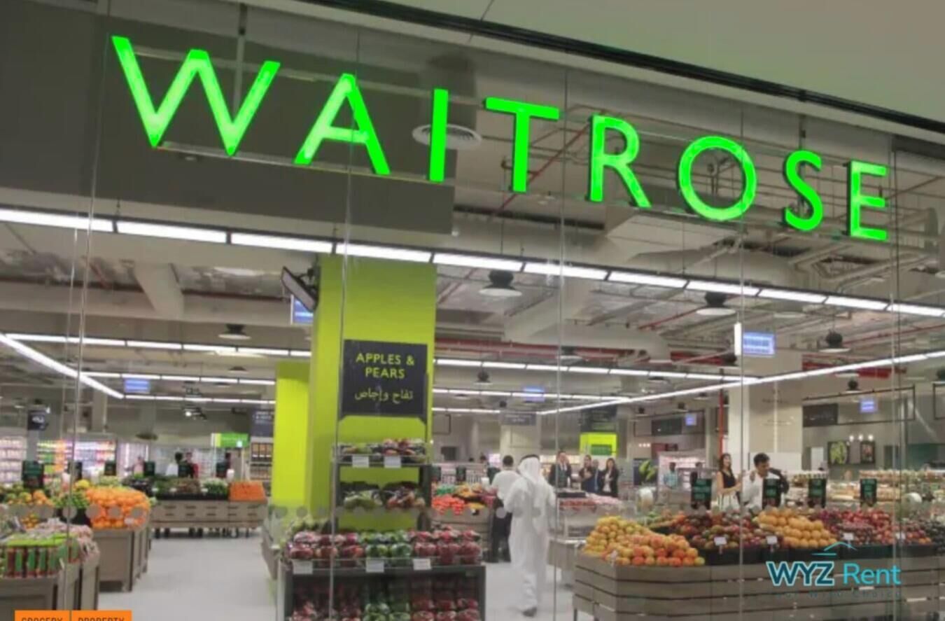 A supermarket interior with a prominent 'WAITROSE' sign, various produce sections, and a visible 'APLES & PEARS' section.