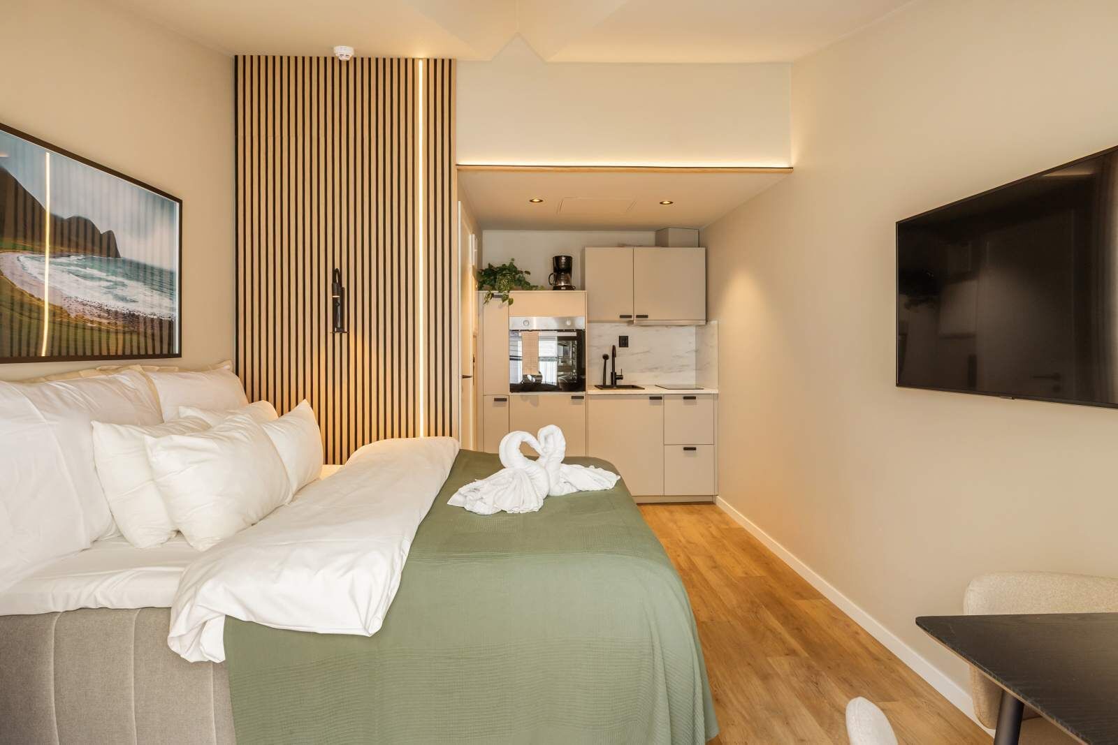 Une chambre d’hôtel avec un lit, une couvre-lit verte, une télévision, un coin cuisine avec des placards et un plan de travail, et un tableau accroché au mur.