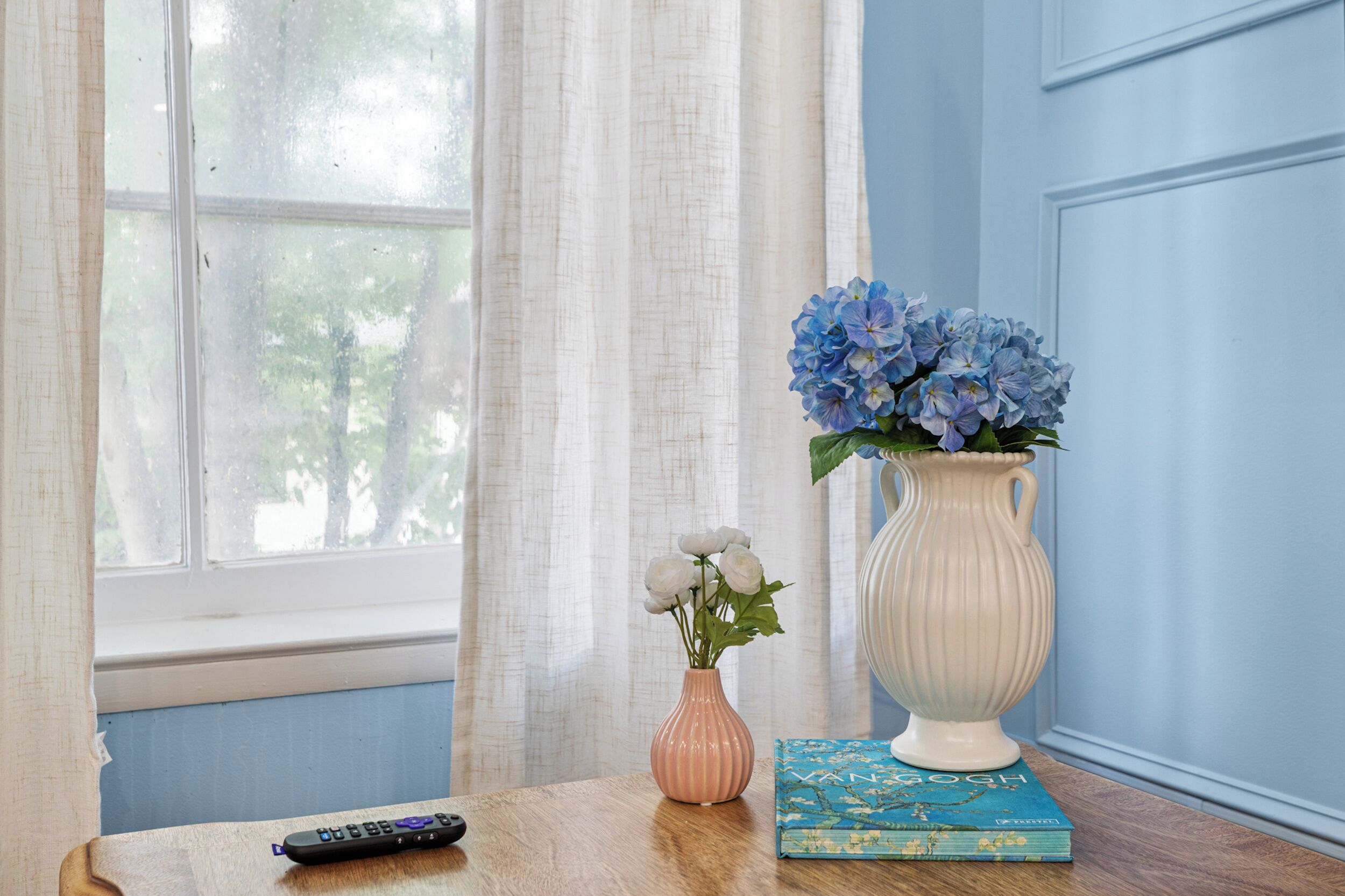 Una habitación con ventana, un jarrón con flores, un control remoto y un libro sobre una mesa.