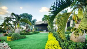 Een verzorgde tuin met palmbomen, strak bijgesnoeide heggen en een bordje met 'RADISIAC BEACH CLUB'.
