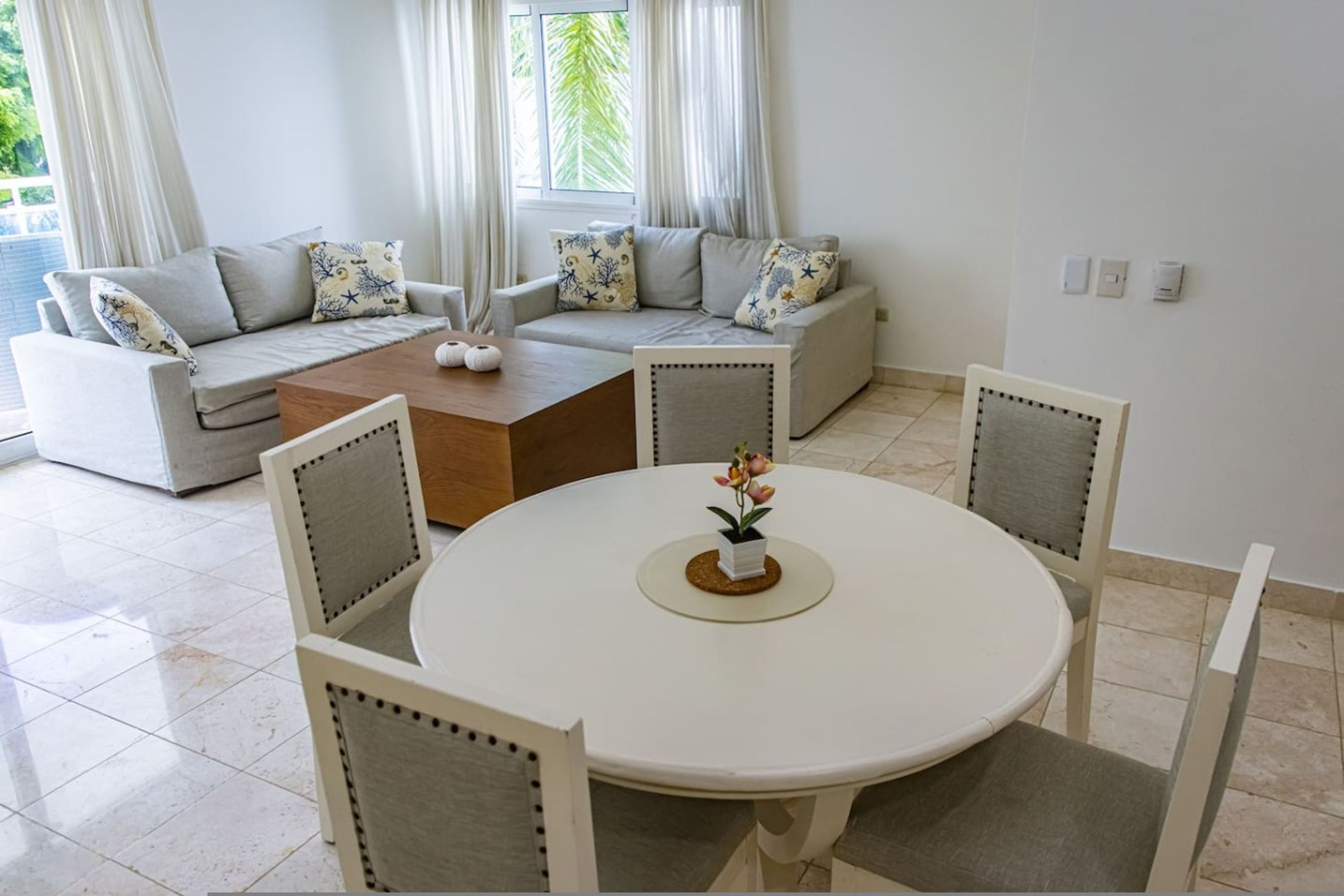 Een moderne woonkamer met een ronde eettafel, grijze banken met gekleurde kussens en een houten salontafel.
