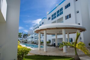 A modern hotel with a pool area, multiple floors, and a covered outdoor seating area. - Beach Condo | 2 Pools | Electricity Included (Punta Cana)