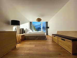 Una camera da letto moderna con un letto, comodini, una scrivania e vista sulle montagne attraverso ampie finestre.