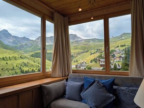 Ein Zimmer mit Holzbalkendecke, einer Fensterbank mit Kissen und Blick auf eine gebirgige Landschaft.