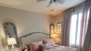 Ein Schlafzimmer mit einem Bett, einem Nachttisch, einem Spiegel, einem Deckenventilator und einem Fenster mit Vorhängen.