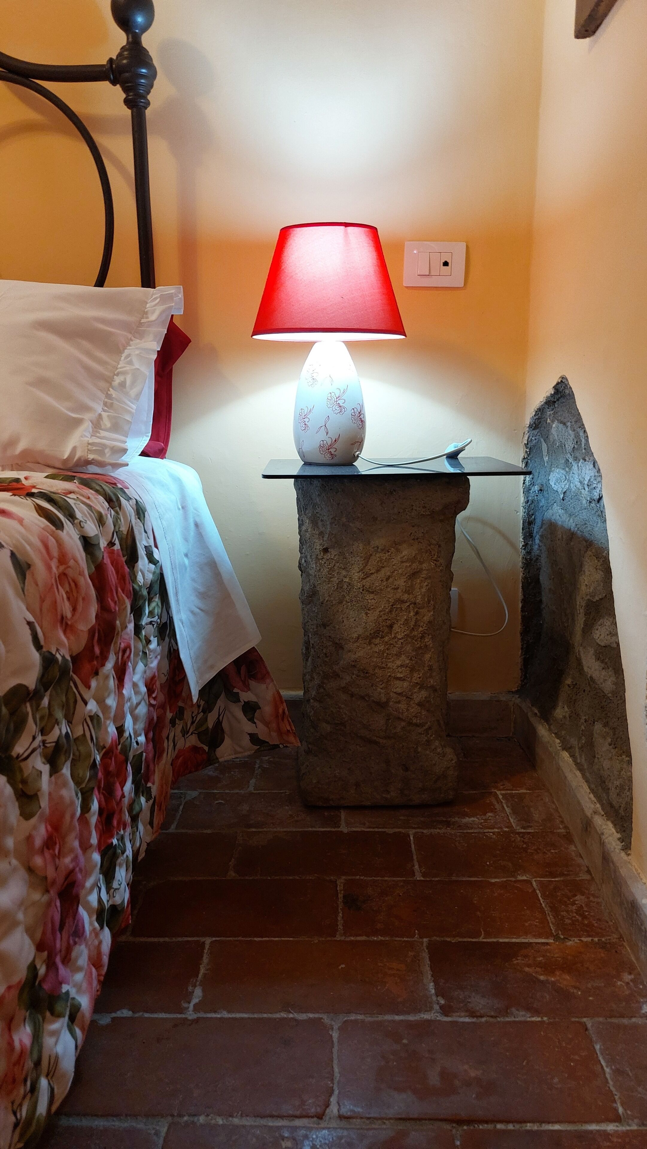 Une chambre avec un lit, une table de chevet en pierre surmontée d’une lampe rouge et un sol carrelé.