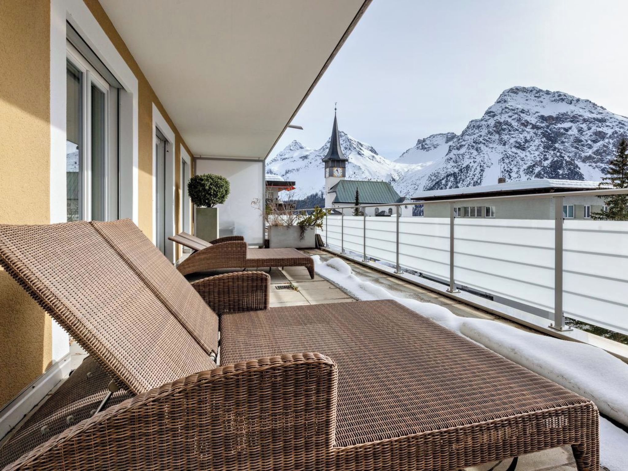 Ein Balkon mit Korbsitzmöbeln, einem Blumentopf und Blick auf schneebedeckte Berge und eine Kirche.