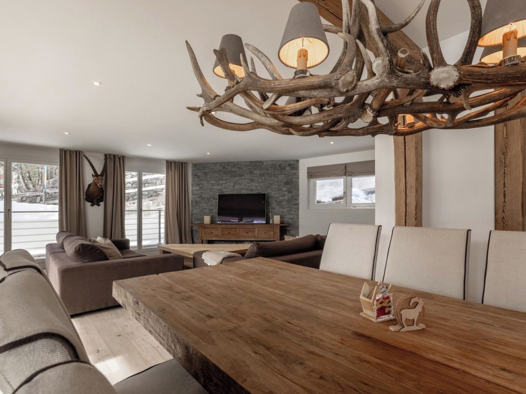 Una sala de estar moderna con una mesa de comedor de madera, sofás de cuero, un televisor montado y una lámpara de araña con cuernos original.