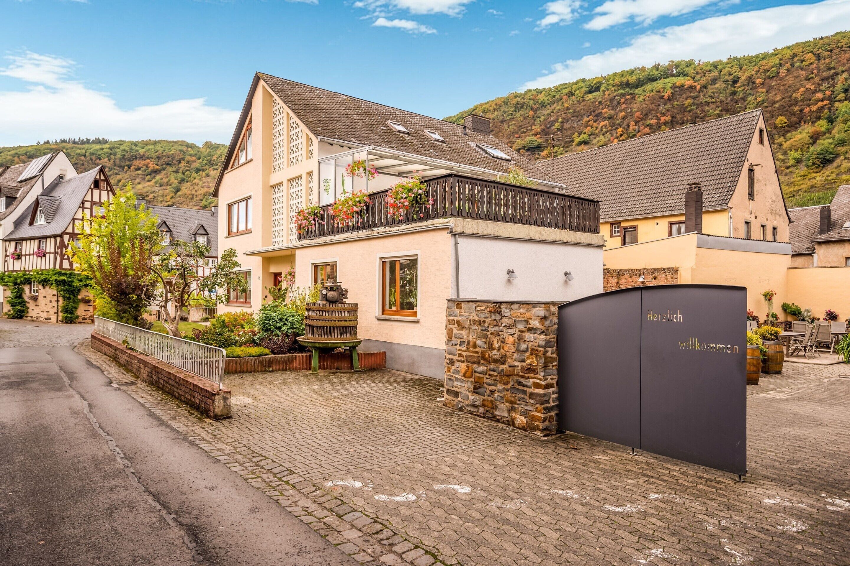 A traditional European house with a stone wall entrance, a balcony with flowers, and a cobblestone driveway.