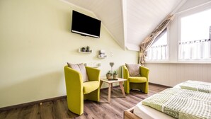 Ein Zimmer mit einem Bett, zwei Sesseln, einem kleinen Tisch, einem an der Wand montierten Fernseher und einem Fenster mit Vorhängen.