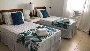 Een hotelkamer met twee bedden, elk voorzien van een sprei met bladmotief, een nachtkastje met een lamp, en een raam met gordijnen.