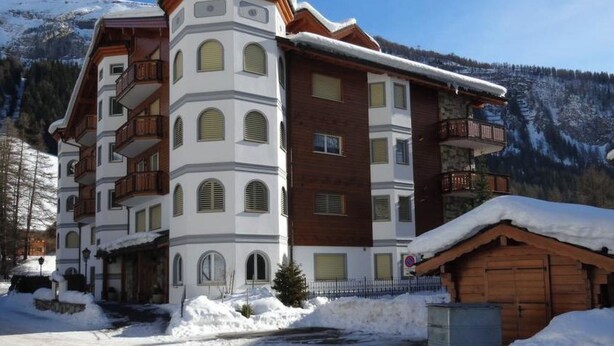 Ein mehrstöckiges Hotel mit schneebedecktem Dach, einem Balkon im oberen Stockwerk und einem Berg im Hintergrund.
