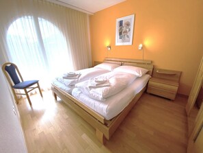 Ein Schlafzimmer mit einem hölzernen Bett, einem Sessel, einem Nachttisch und einem Fenster mit Vorhängen.
