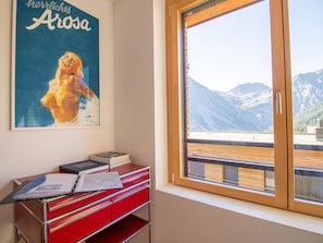 Ein Zimmer mit großem Fenster und Blick auf Berge, ein roter Schreibtisch mit Büchern und ein Poster an der Wand.