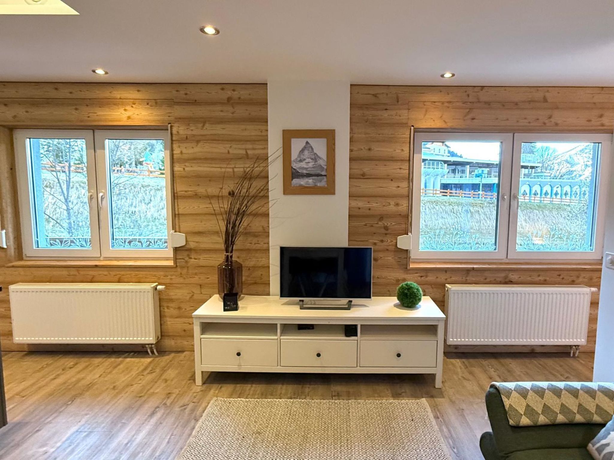 A modern living room with wooden panel walls, a flat-screen TV, a white entertainment unit, and large windows with a view of a building and trees.
