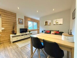 A modern living room with a wooden dining table, black chairs, a flat-screen TV, and a window with a view of trees. - Chalet Lucia 1 (EG) # 3-bed apartment, Dusche, WC 3-bed apartment, Dusche, WC by Interhome (Leukerbad)