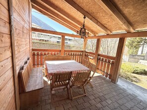 A wooden deck with a table and chairs, a hanging light fixture, and a view of a building and trees. - Chalet Lucia 1 (EG) # 3-bed apartment, Dusche, WC 3-bed apartment, Dusche, WC by Interhome (Leukerbad)