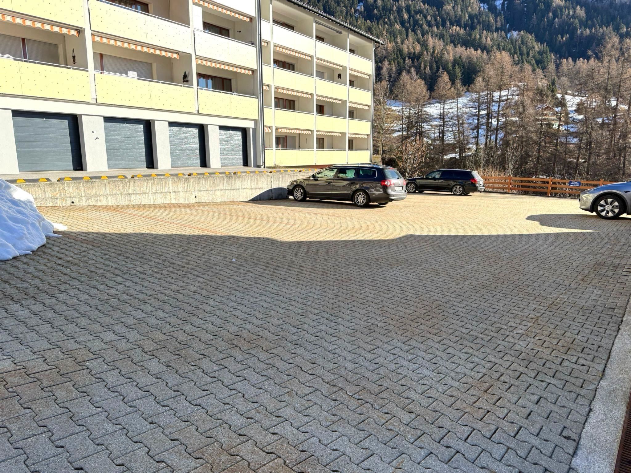 Un estacionamiento con vehículos, un edificio de varios pisos y balcones, y montañas al fondo.