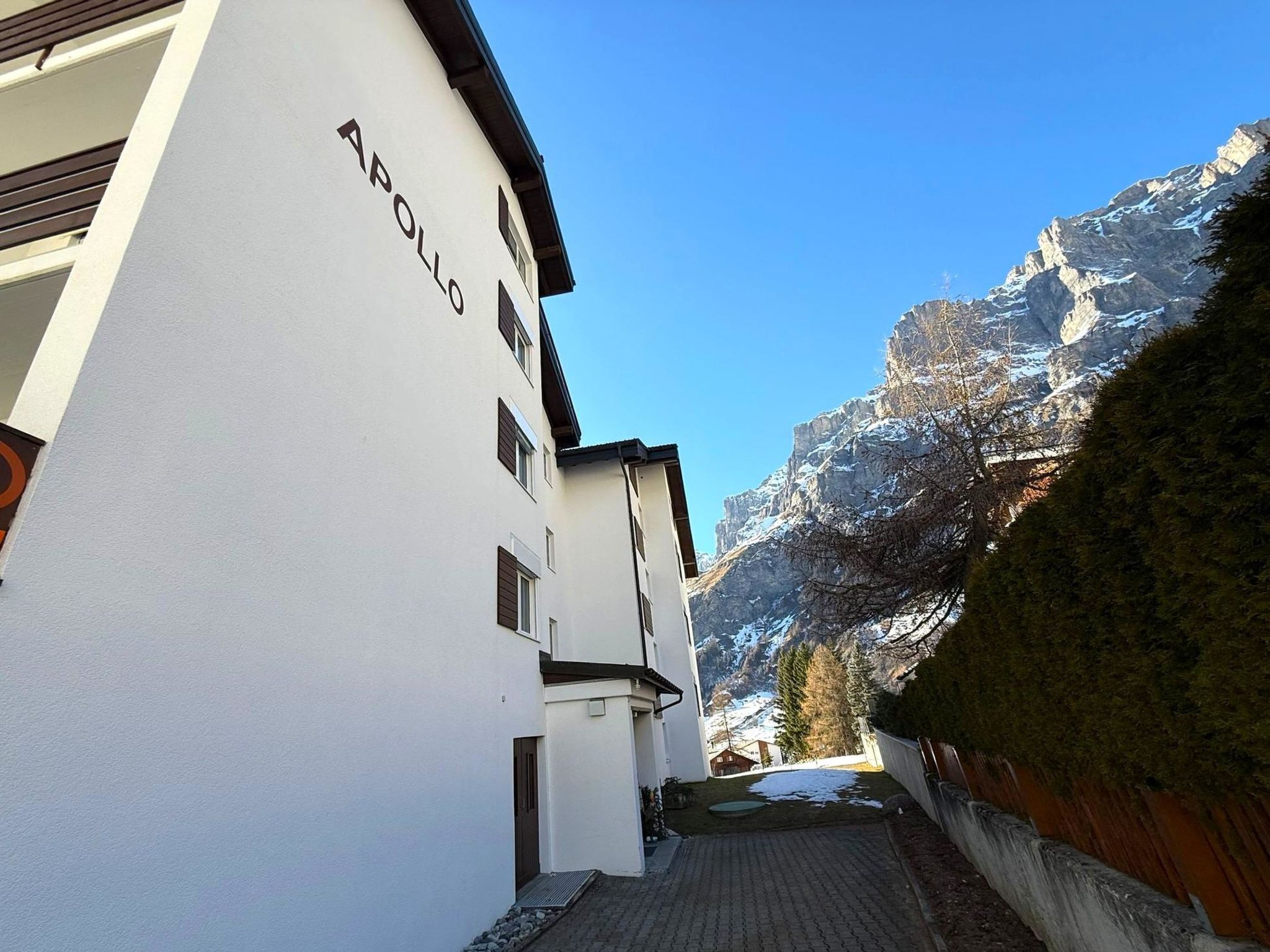 Eine schmale Straße mit weißen Gebäuden, eines mit der Aufschrift 'APOLLO', die zu einer Bergblick führt.