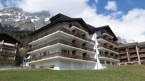 Een hotel met meerdere verdiepingen, balkons, een gazon op de voorgrond en een bergachtig landschap op de achtergrond.