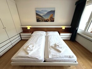 A double bed with white linens and towels, two bedside tables with lamps, a painting of a mountain landscape on the wall, and a window with curtains. - Apollo 50 # 4-bed apartment, Bad, WC 4-Bettwohnung, Bad, WC by Interhome (Leukerbad)