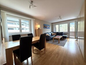 A spacious living area with a dining table, chairs, a sofa, and a coffee table. There are large windows providing natural light and views of the outside. - Apollo 50 # 4-bed apartment, Bad, WC 4-Bettwohnung, Bad, WC by Interhome (Leukerbad)