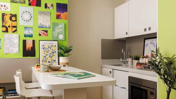 Una cocina moderna con encimera blanca, un lavamanos y un microondas. Hay cuadros en la pared, una planta en maceta y una pequeña mesa con un cuenco y un libro.
