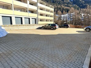 A parking area with vehicles, a building with multiple floors and balconies, and a mountainous backdrop. - Apollo 47 # 2-bed apartment, Dusche, WC 2-Bettwohnung, Dusche, WC by Interhome (Leukerbad)