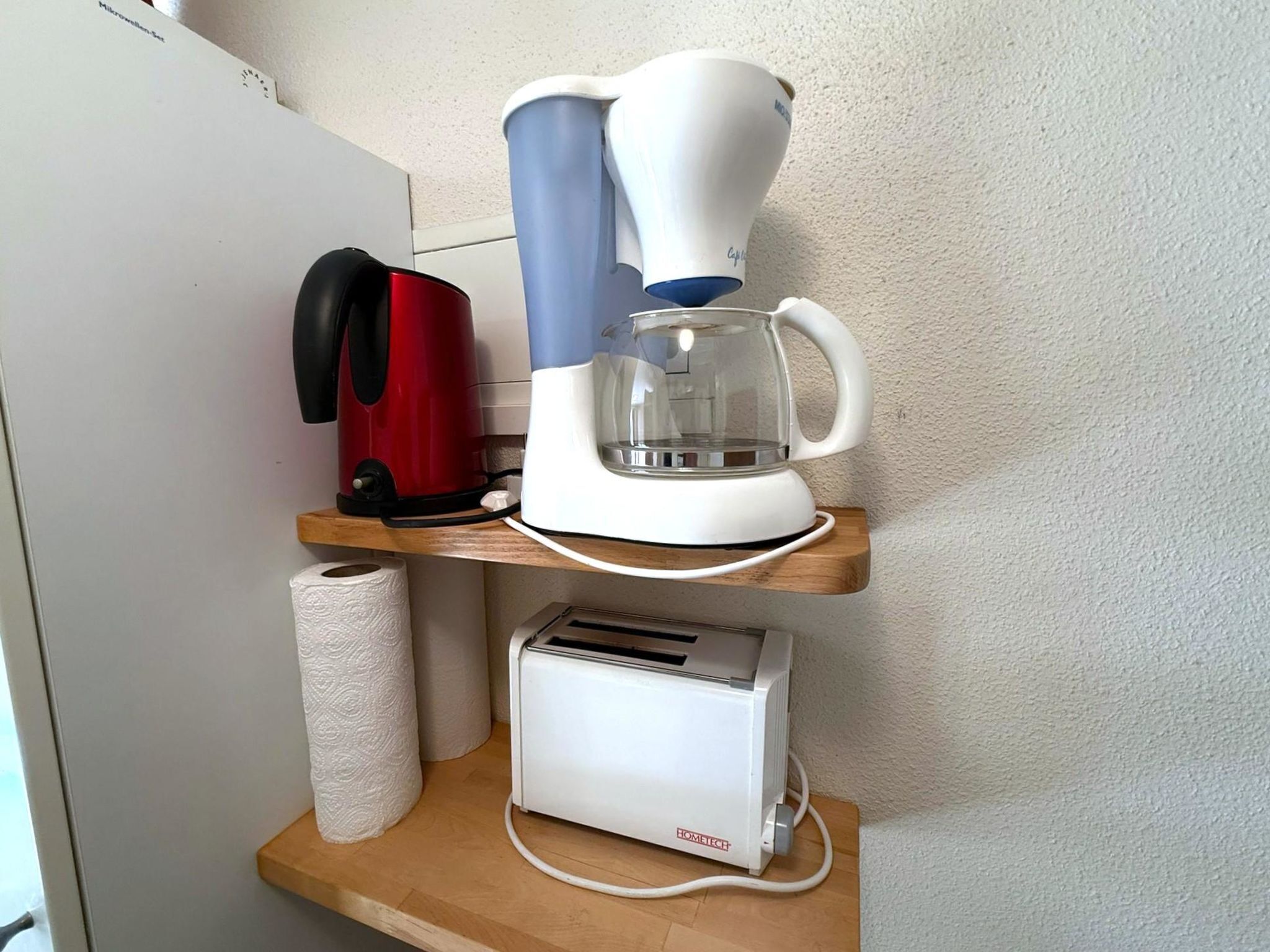 A kitchenette with a coffee maker, toaster, and a roll of paper towels on a wooden shelf.