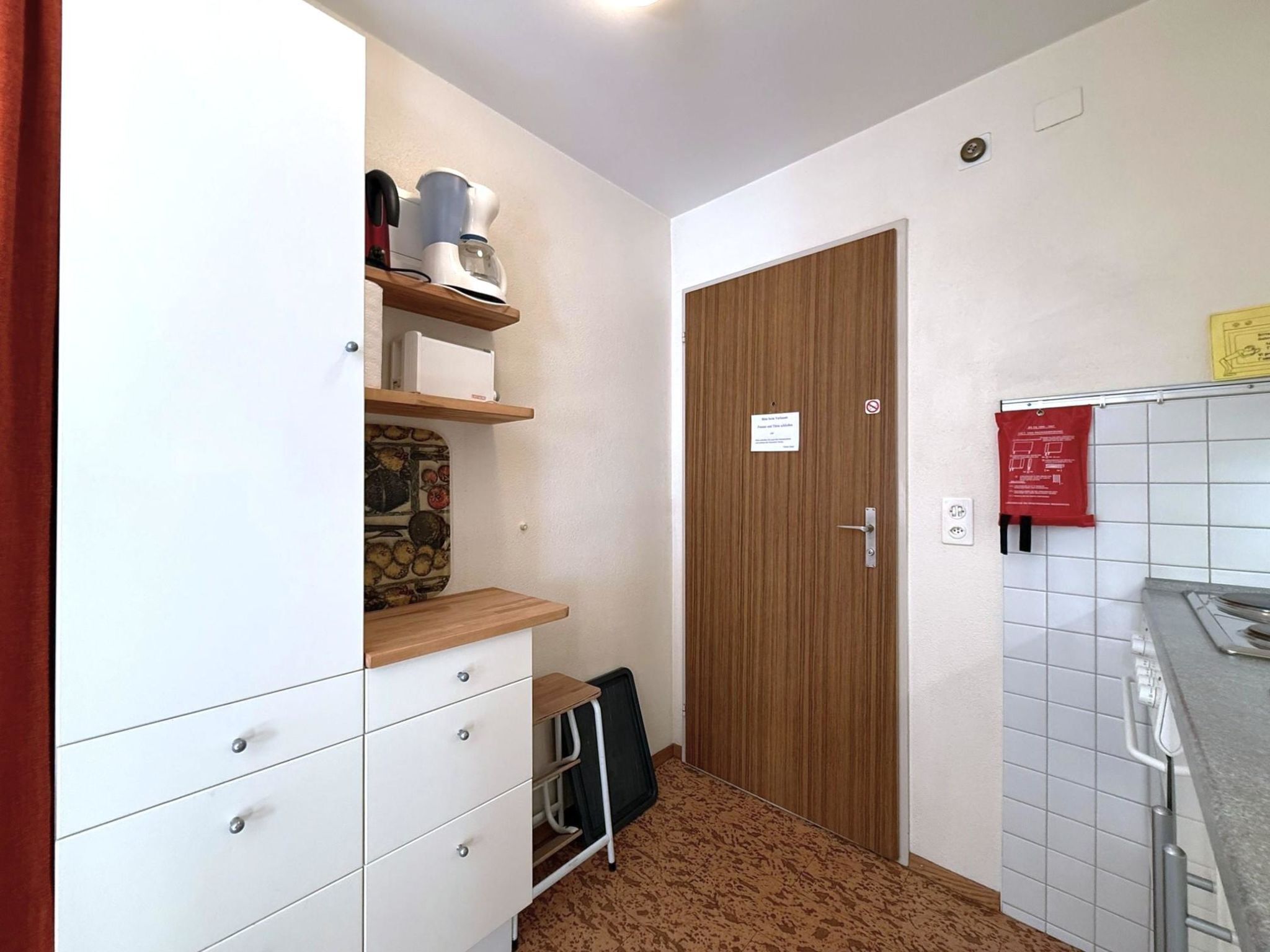 Une petite cuisine équipée avec une étagère en bois, un placard blanc et une porte sur laquelle est apposée une pancarte.