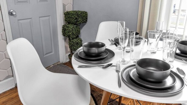Una mesa de comedor dispuesta con platos, tazones y cubiertos negros, rodeada de sillas blancas, con una planta pequeña en maceta y una puerta al fondo.