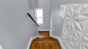 Eine enge Treppe mit Holzdielen führt zu einem Podest, ein Fenster mit Jalousien und eine Wand mit geometrischem Muster.