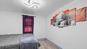 Un dormitorio con una cama, una ventana con cortinas moradas, una lámpara de pared y una serie de cuadros que representan cascadas.