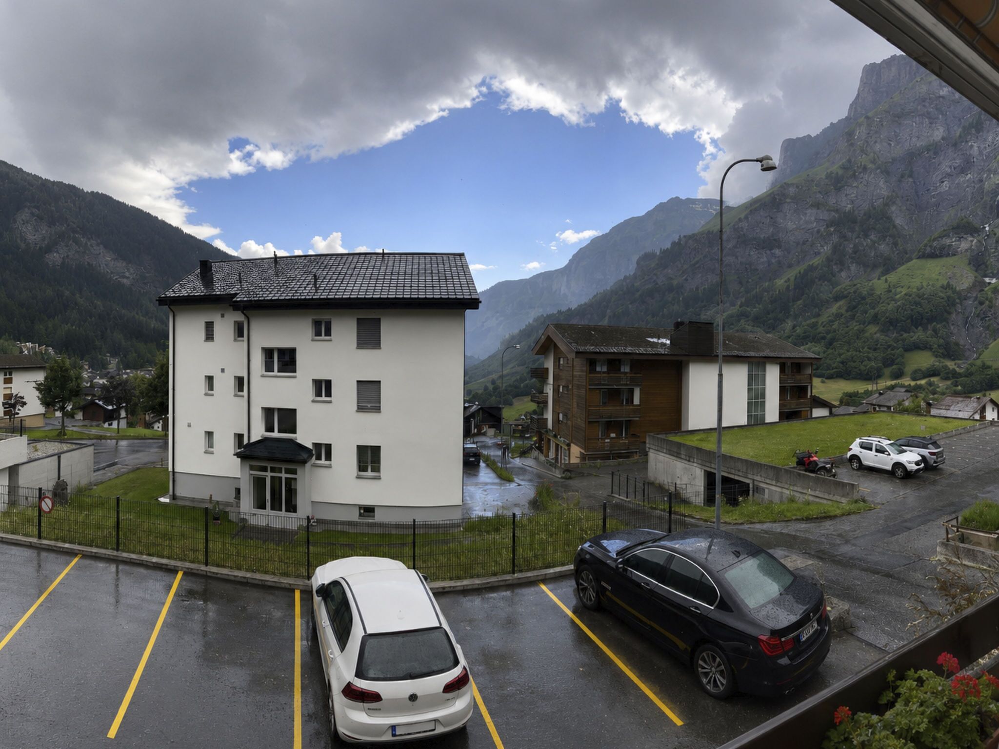 Ein mehrgeschossiges Gebäude mit flachem Dach, umgeben von einem Parkplatz mit Autos, und eine gebirgige Landschaft im Hintergrund.