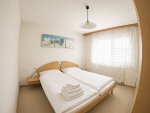 Ein Schlafzimmer mit einem Doppelbett, einem Nachttisch mit Lampe, einem Fenster mit durchsichtigen Vorhängen und zwei an der Wand hängenden Bildern.