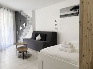 Ein modernes Schlafzimmer mit grauem Sofa, einem kleinen runden Tisch, einem Bett mit weißer Bettwäsche und einer Wandleuchte.