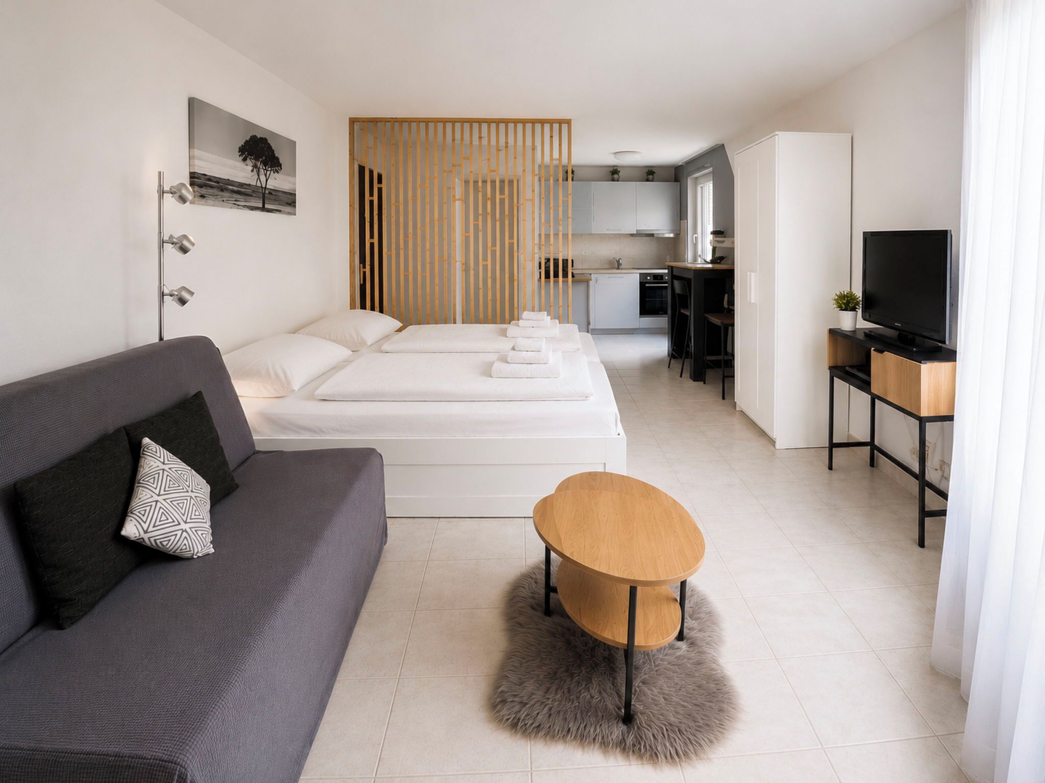 A modern hotel room with a grey sofa, a wooden coffee table, a bed with white linens, a kitchen area, and a television.