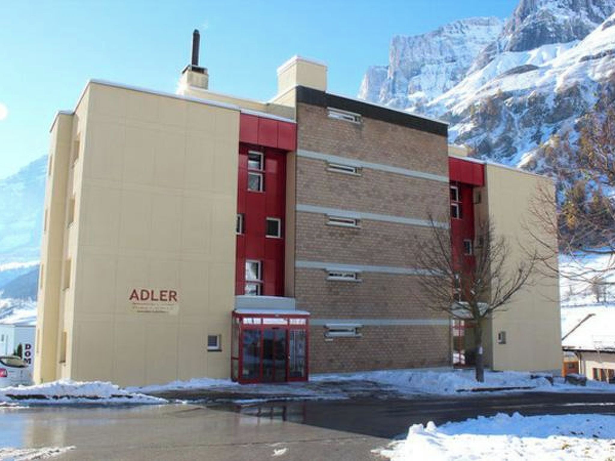 A building with the name 'ADLER' on the facade, a red entrance, and a snow-covered mountain in the background.