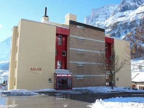 A building with the name 'ADLER' on the facade, a red entrance, and a snow-covered mountain in the background. - Adler 82 # 2-bed apartment, Dusche, WC 2-Bettwohnung, Dusche, WC by Interhome (Leukerbad)