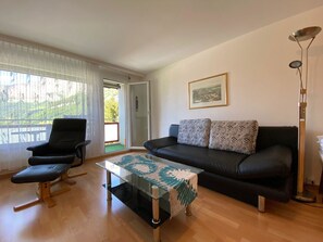 Ein Wohnzimmer mit einem schwarzen Ledersofa, einem schwarzen Ledersessel, einem Glastisch mit Läuferdecke mit Blattmuster und einer Stehlampe.